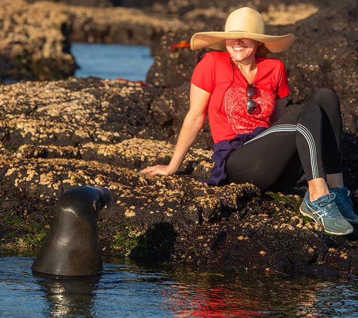 Woman wearing a sun hat sitting on the rocky shore next to a Galapagos Sea Lion