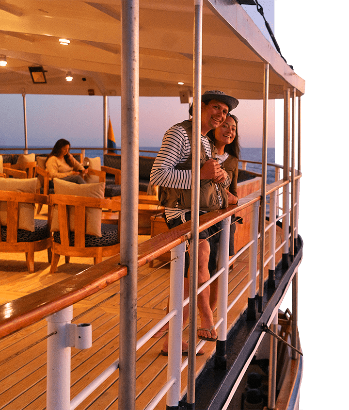 Couple leaning on the Grace yacht top deck rail, looking off into the sunset